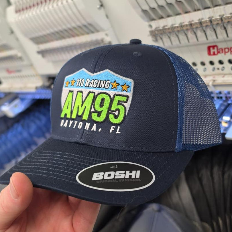 Navy blue cap with racing-themed design held in front of embroidery equipment