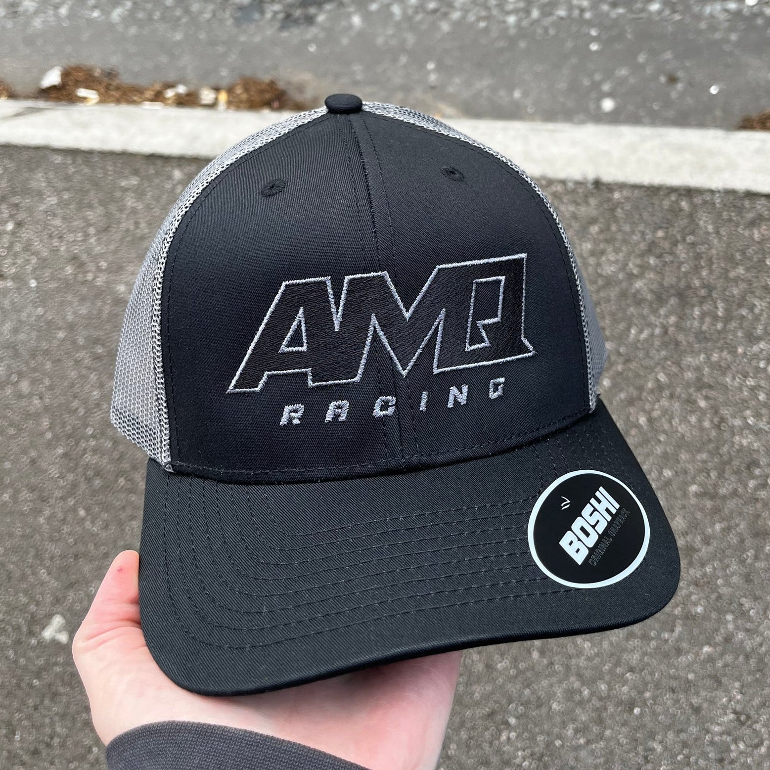 Black and gray cap with AMR Racing logo held by a hand on a road background