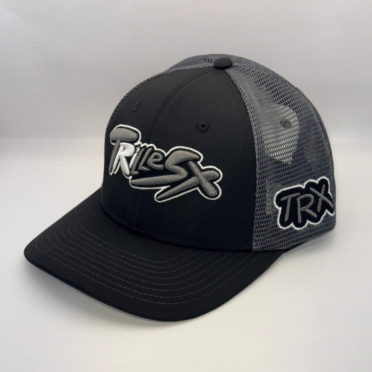 A black and gray trucker hat with a curved brim, featuring a black front panel with a stylized gray and white 'Trix' logo, black mesh back panels, and a black 'TRX' logo on the side.