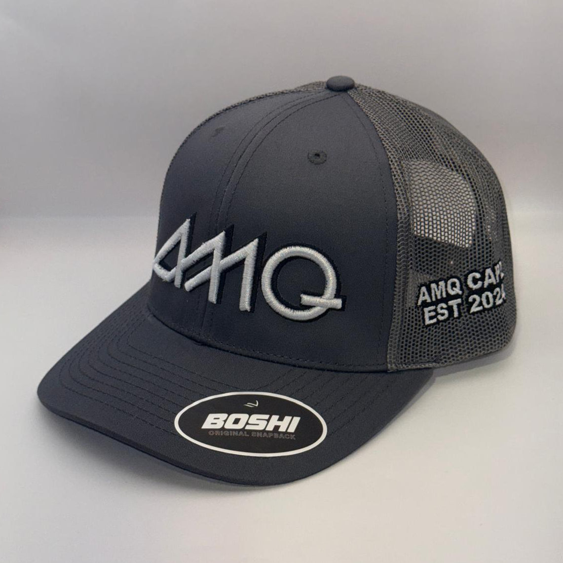 A dark gray AMQ trucker hat with a curved brim, featuring a white embroidered AMQ logo on the front, white text on the side mesh, and a circular white BOSHI logo patch on the brim.