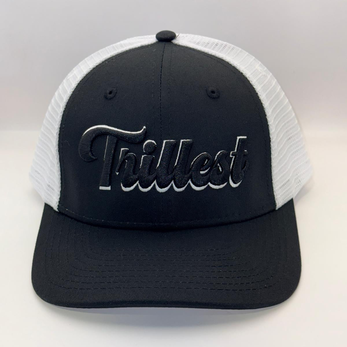 A black and white trucker hat with a black front panel, white mesh back, and a black embroidered 'Trilkest' logo on the front.