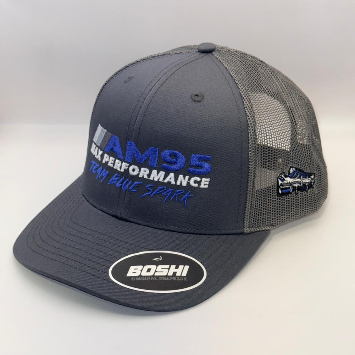 A dark gray and black mesh-back trucker hat with 'AM95 ALK PERFORMANCE' embroidered on the front in blue and white, featuring a white oval 'BOSHI' sticker on the brim and a small side patch.
