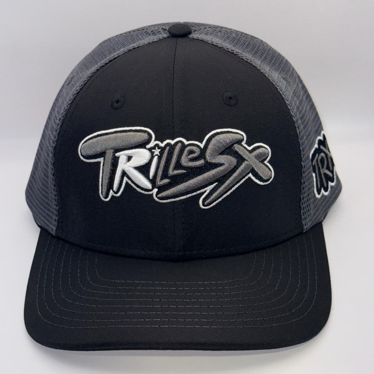 A black and gray trucker hat with a black front panel, gray mesh back, and a large stylized 'TRILEX' logo embroidered on the front.