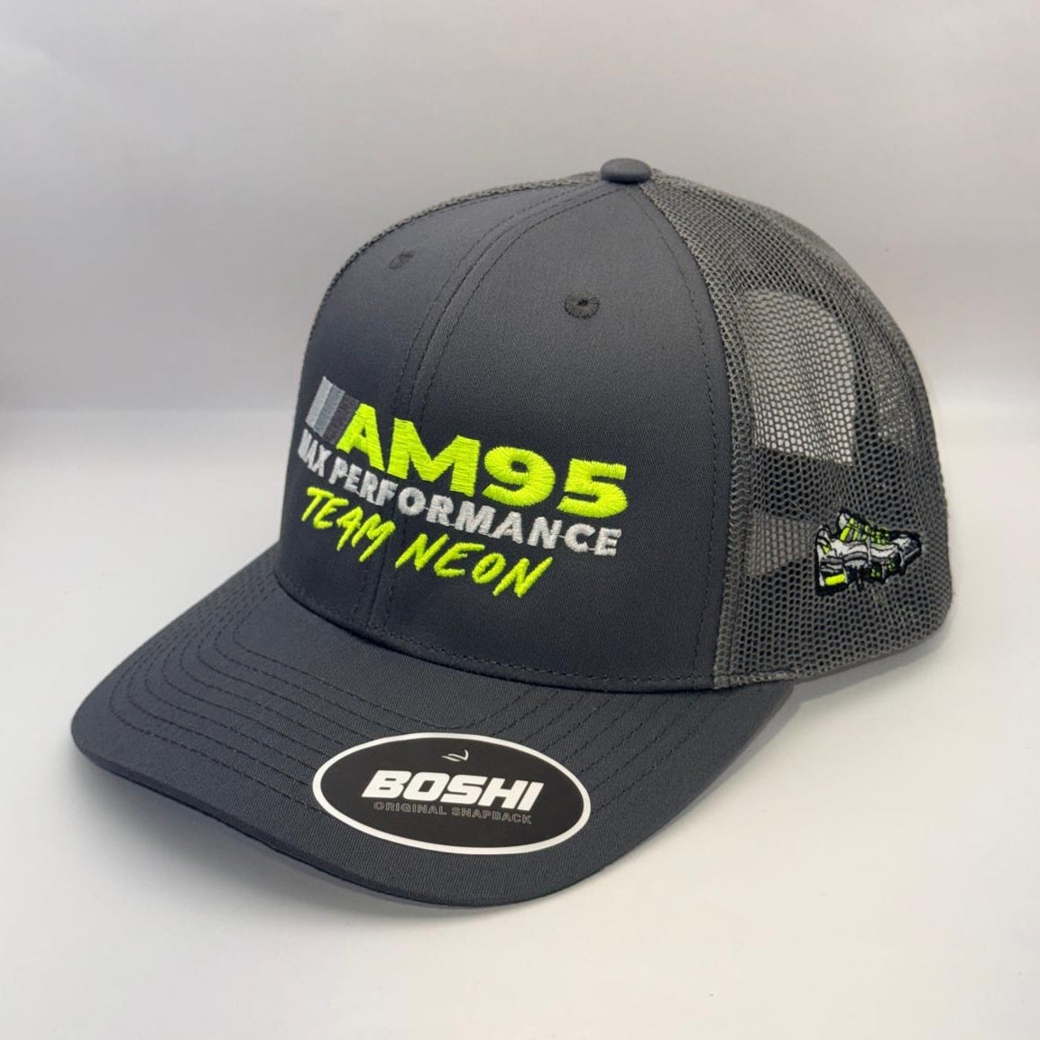 A dark gray mesh-back trucker hat with 'AM95 MX PERFORMANCE TEAM NEON' printed on the front in neon yellow and white, featuring a white oval 'BOSHI' sticker on the brim and a small side patch.