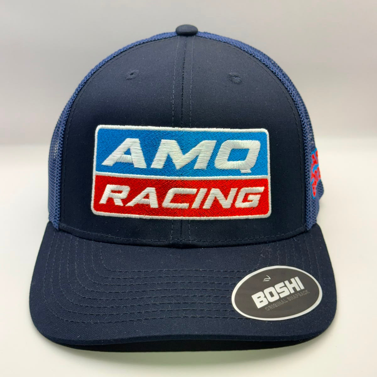 A navy blue trucker-style cap with a large front patch reading 'AMQ RACING' in white, blue, and red. The cap has navy mesh back panels and a navy curved brim with a visible 'BOSHI' sticker on the side.