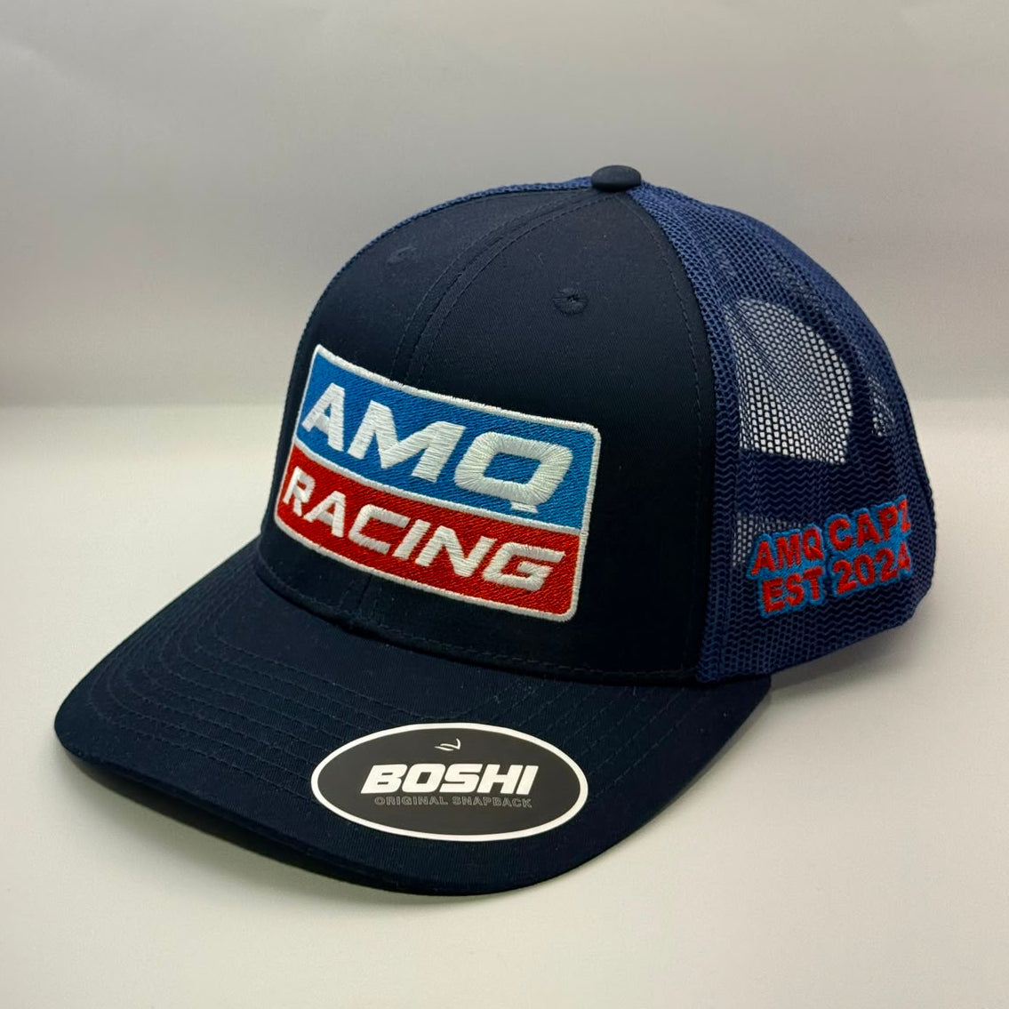 A navy blue and navy mesh trucker hat featuring a prominent 'AMQ RACING' embroidered patch on the front and a 'BOSHI' sticker on the visor, set against a plain light gray background.