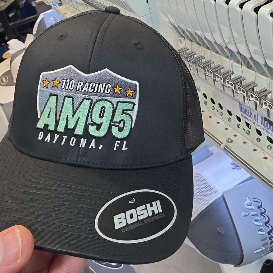Black cap with racing logo and text held in front of embroidery machines