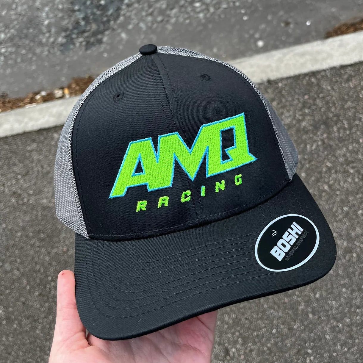 Black and gray cap with green 'AMQ Racing' logo held by a hand on a road surface.